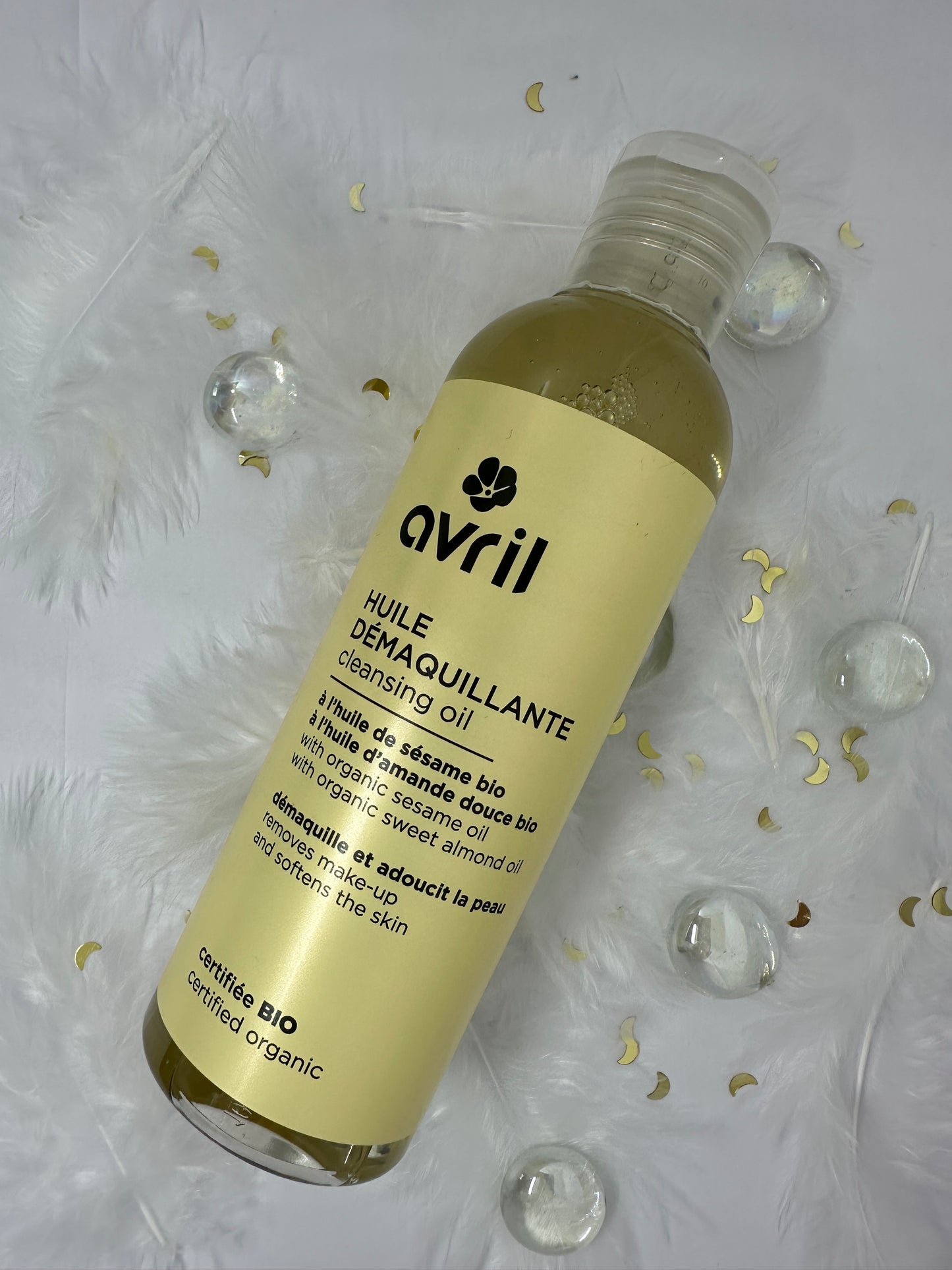 huile démaquillante avril sur fond blanc avec plumes blanches, paillettes dorées et billes transparentes