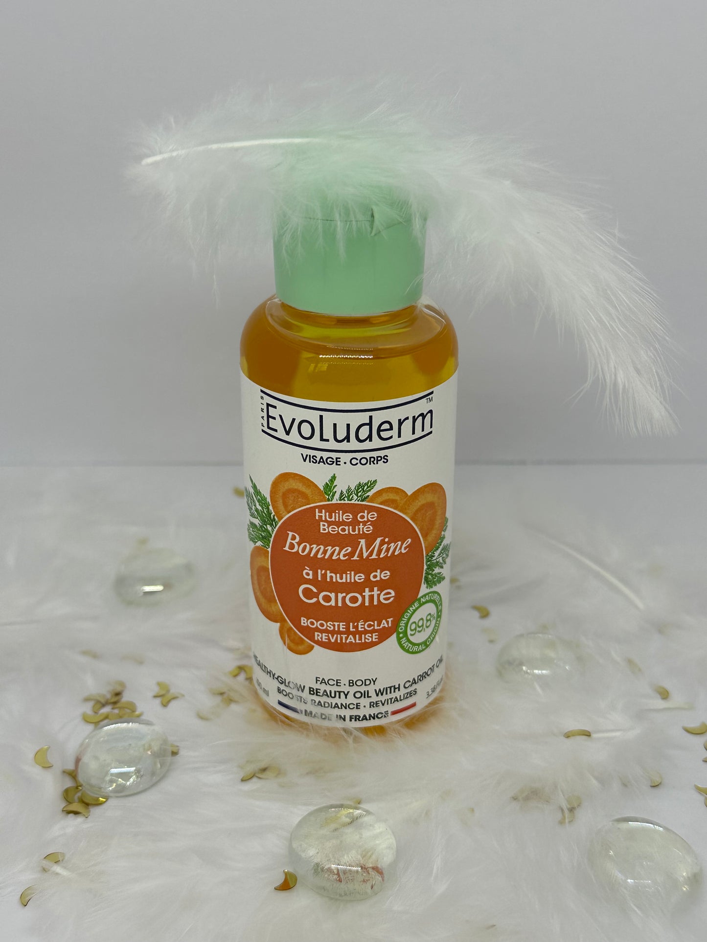 Huile bonne mine à la carotte Evoluderm sur décor blanc avec paillettes, plumes  et billes transparentes - soin éclat du Coffret Teint Radieux