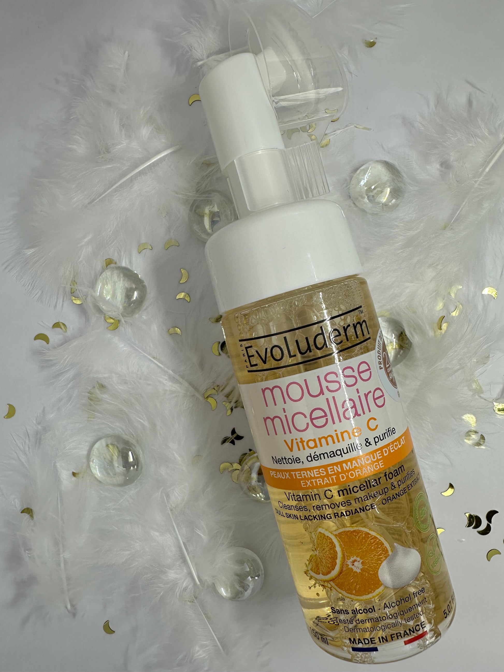 Mousse micellaire Evoluderm vitamine C sur décor blanc, paillettes, plumes et billes plates transparentes - nettoyant visage doux 150 ml