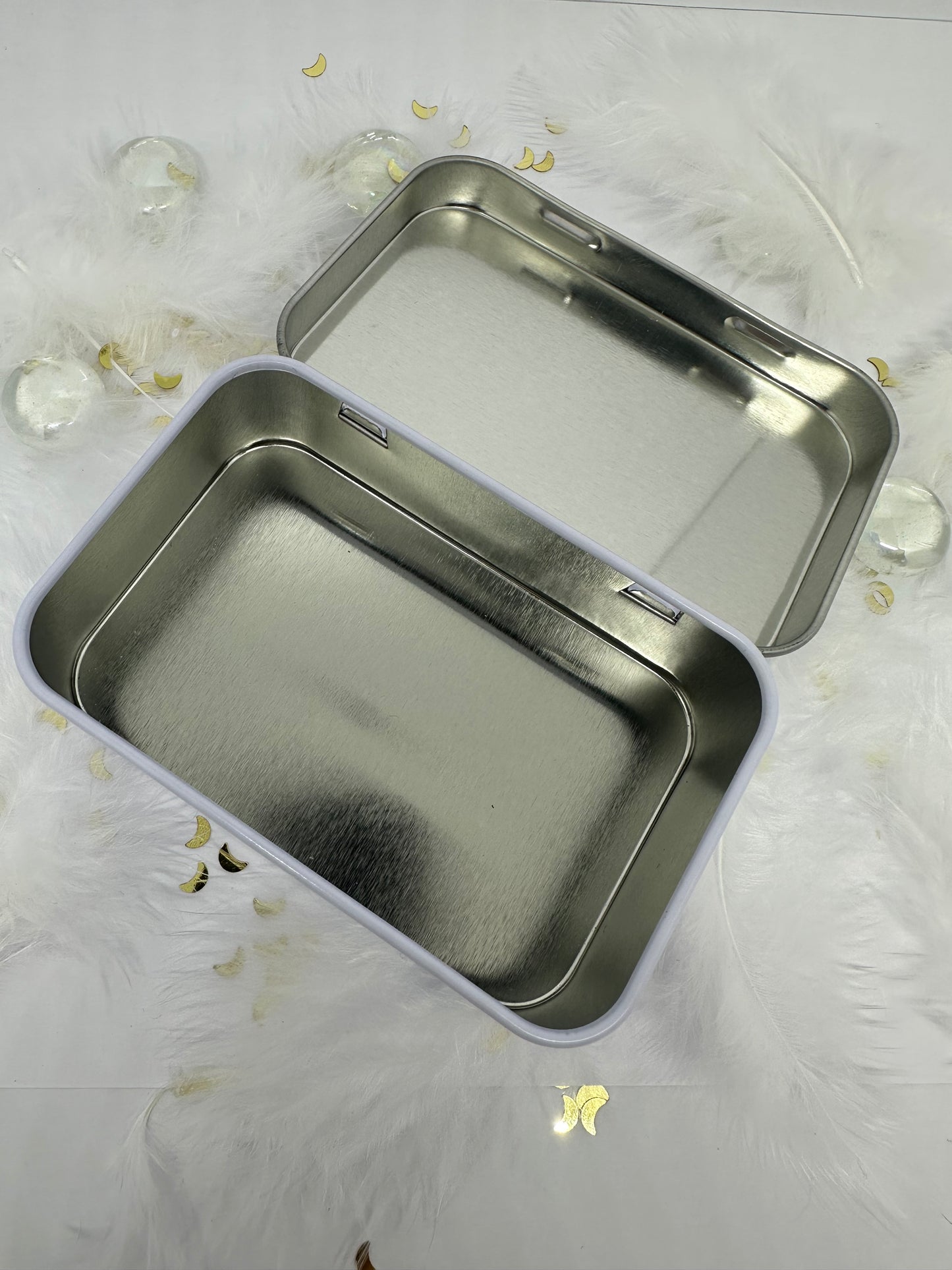boite à savon métal blanc ouverte sur fond blanc avec plumes blanches, paillettes dorées et billes transparentes