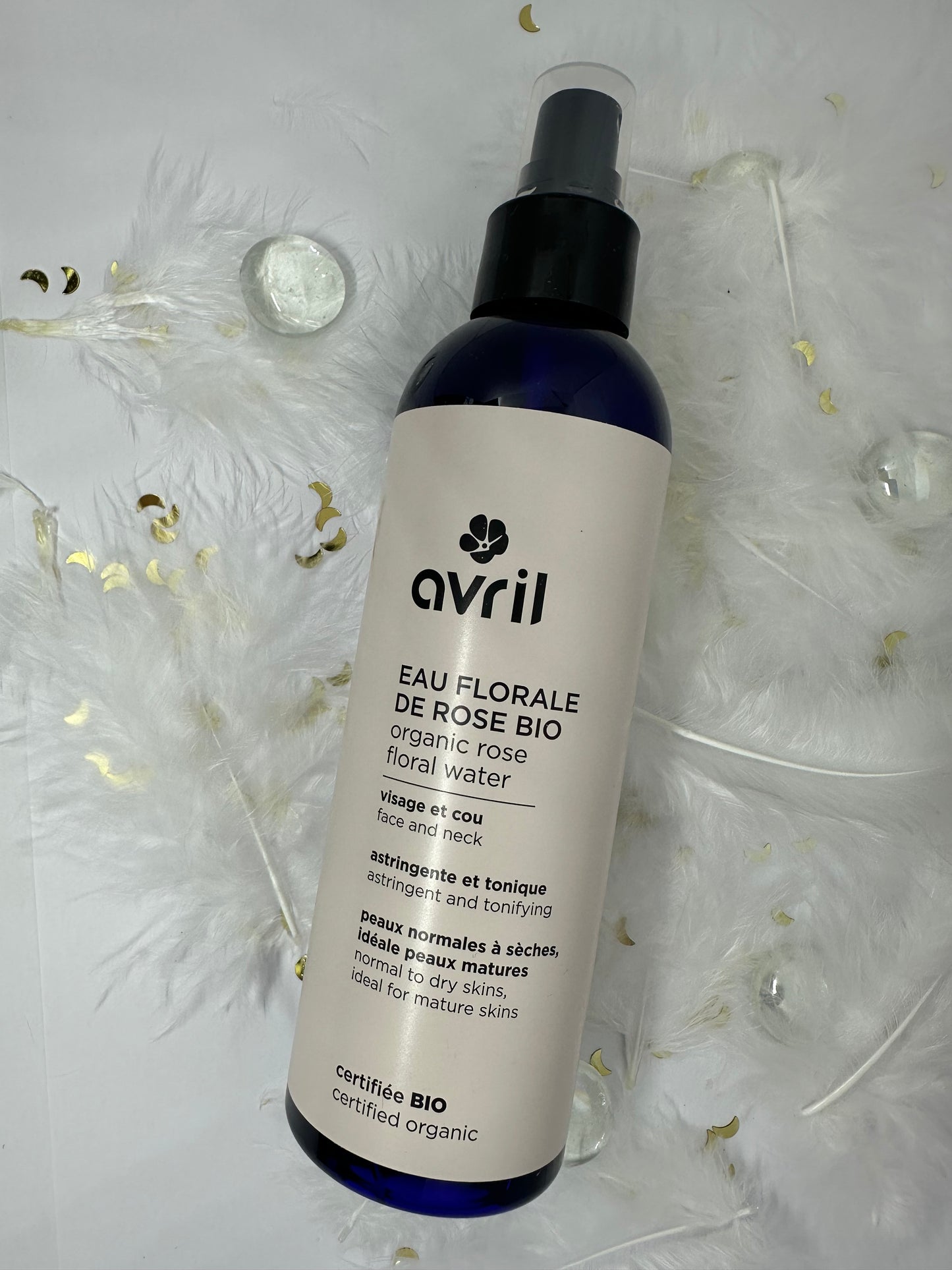 eau florale de rose bio avril sur fond blanc avec plumes blanches, billes transparentes et paillettes dorées