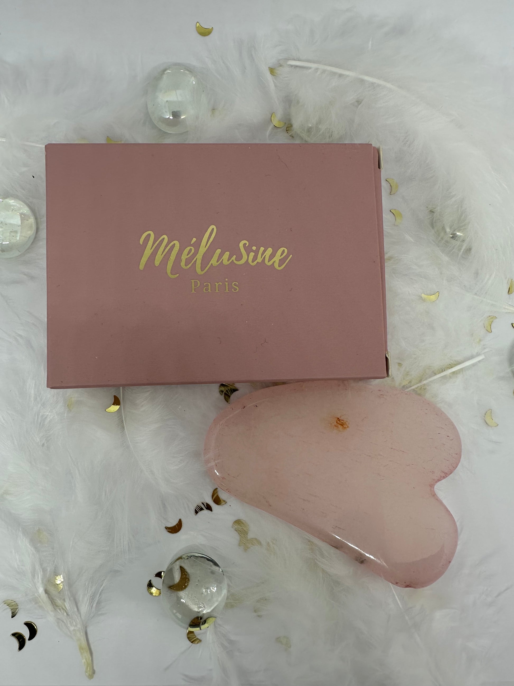 Gua sha quart rose et son emballage carton rose sur fond blanc avec plumes blanches, paillettes dorées et  billes transparentes