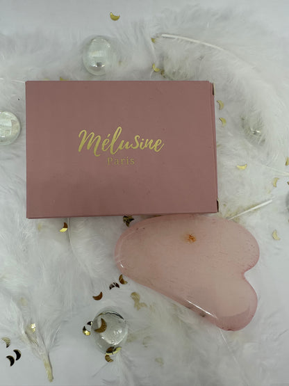 Gua sha quart rose et son emballage carton rose sur fond blanc avec plumes blanches, paillettes dorées et  billes transparentes