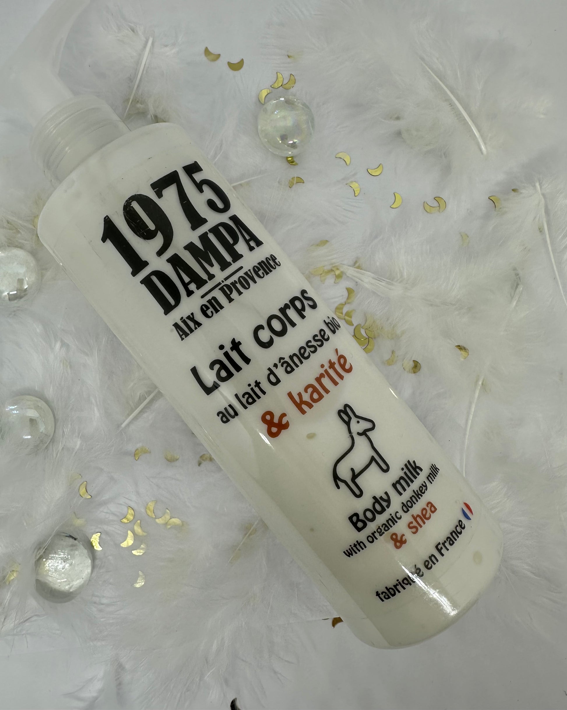 lait corps anesse et karité lanc sur fond blanc avec plumes blanches, paillettes dorées et billes transparentes
