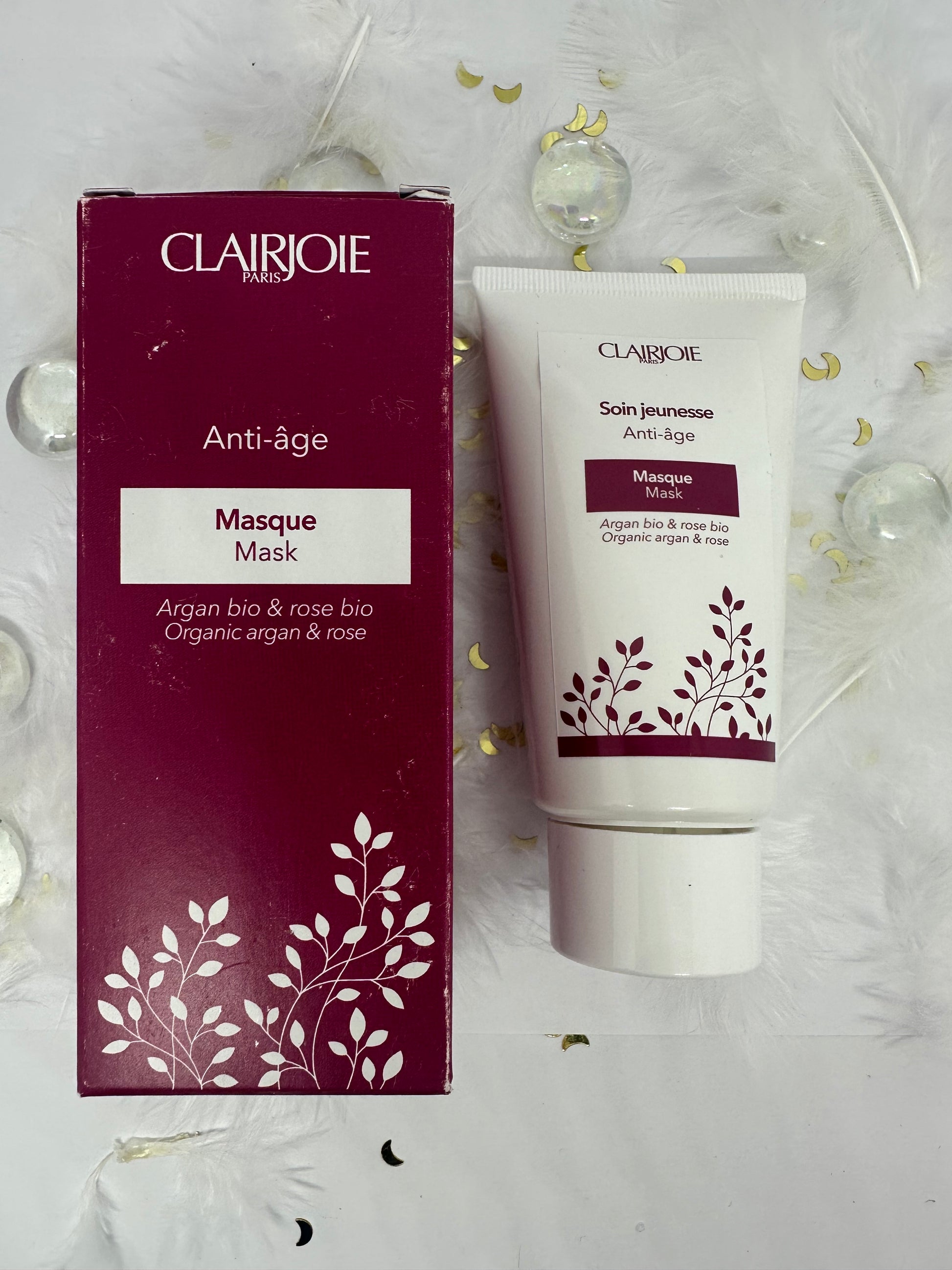 masque clairjoie blanc avec son emballage bordeaux sur fond blanc avec plumes blanches, paillettes dorées et billes transparentes
