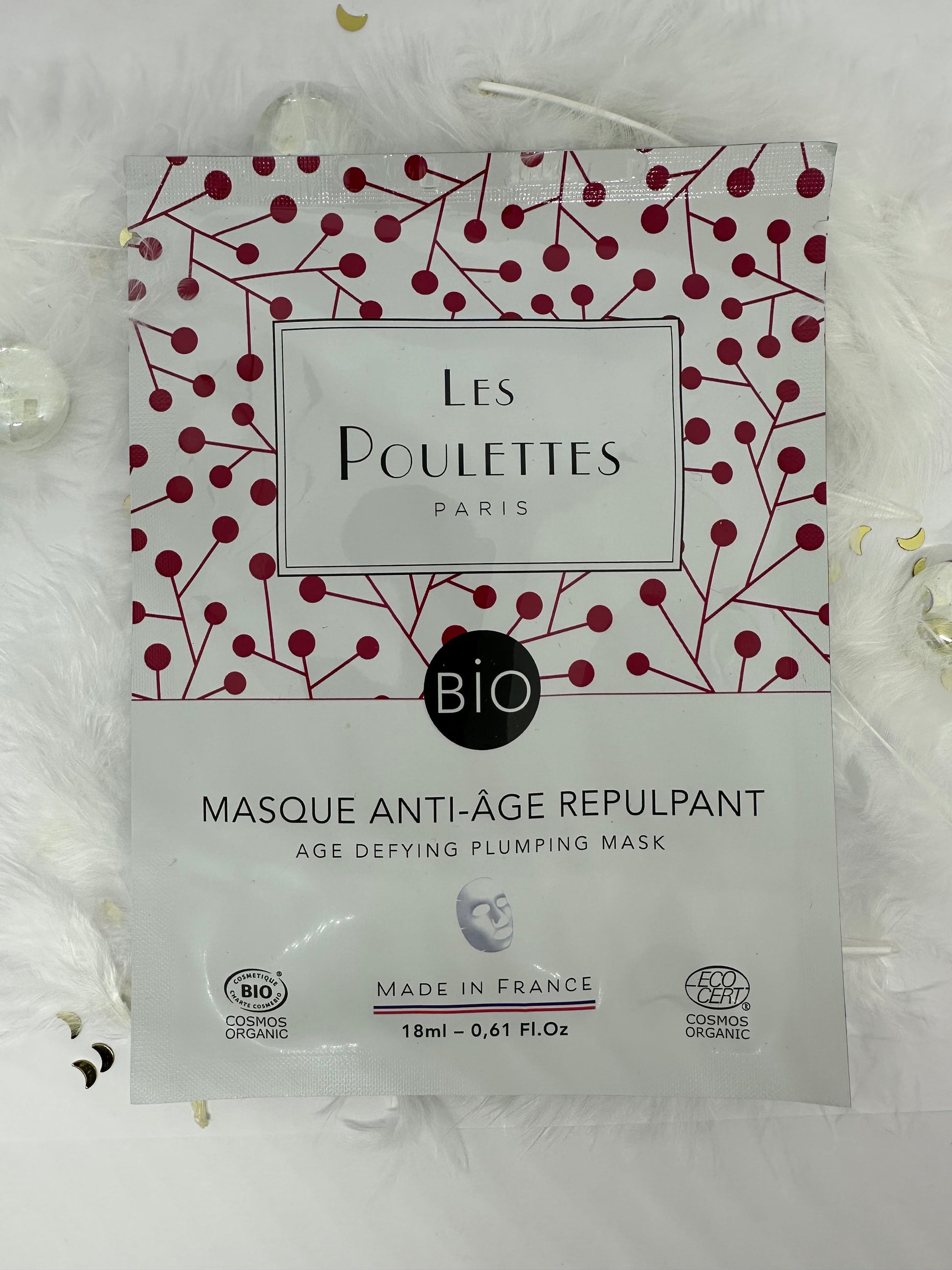 Masque anti-âge repulpant Les Poulettes Paris en sachet blanc, photographié sur fond blanc avec plumes, paillettes dorées et billes transparentes