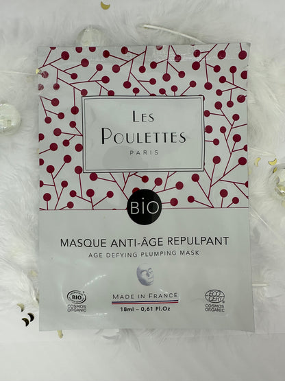 Masque anti-âge repulpant Les Poulettes Paris en sachet blanc, photographié sur fond blanc avec plumes, paillettes dorées et billes transparentes
