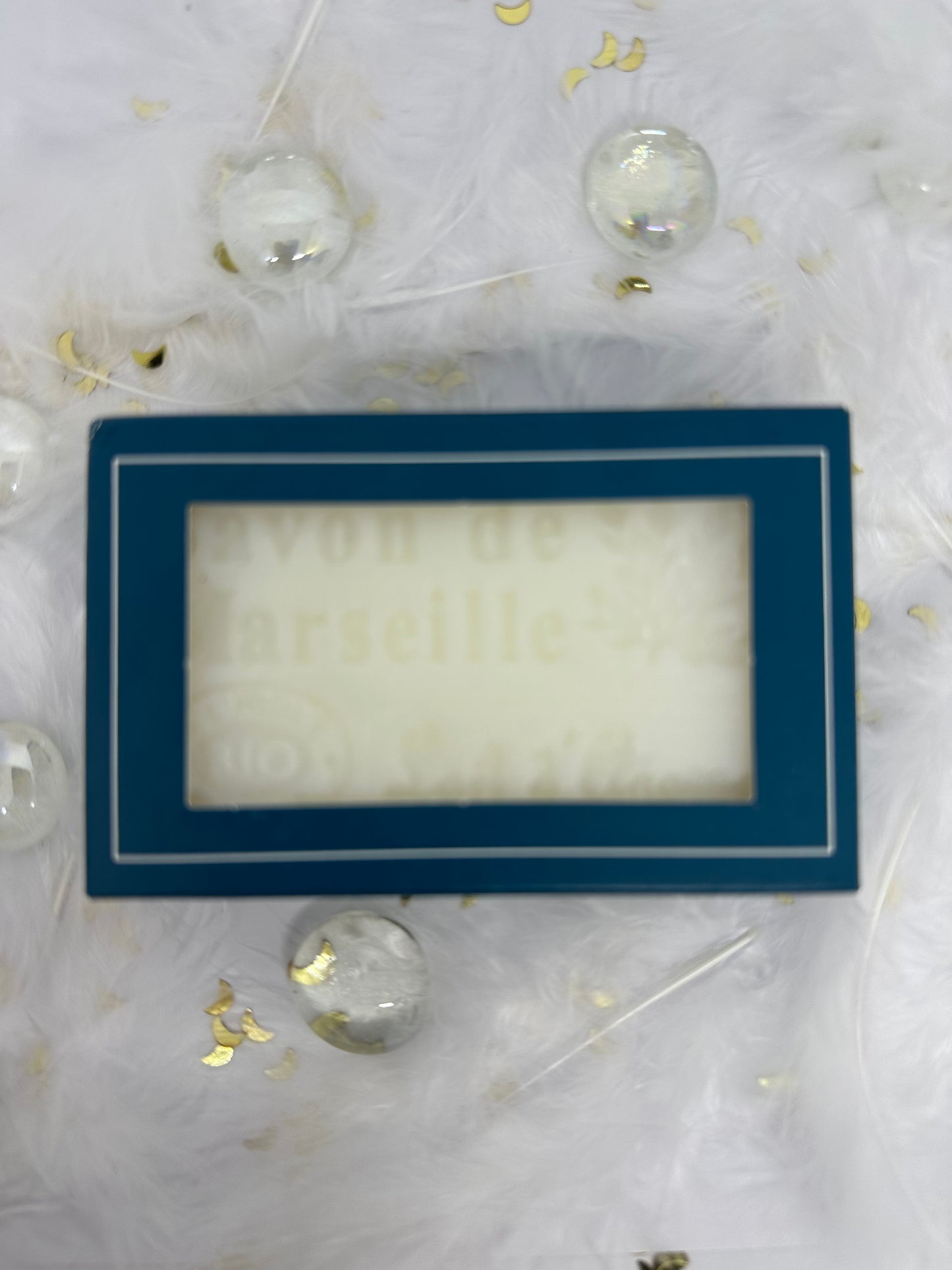 Savon de marseille blanc dans son emballage carton bleu sur fond blanc avec plumes blanches, paillettes dorée et billes transparentes