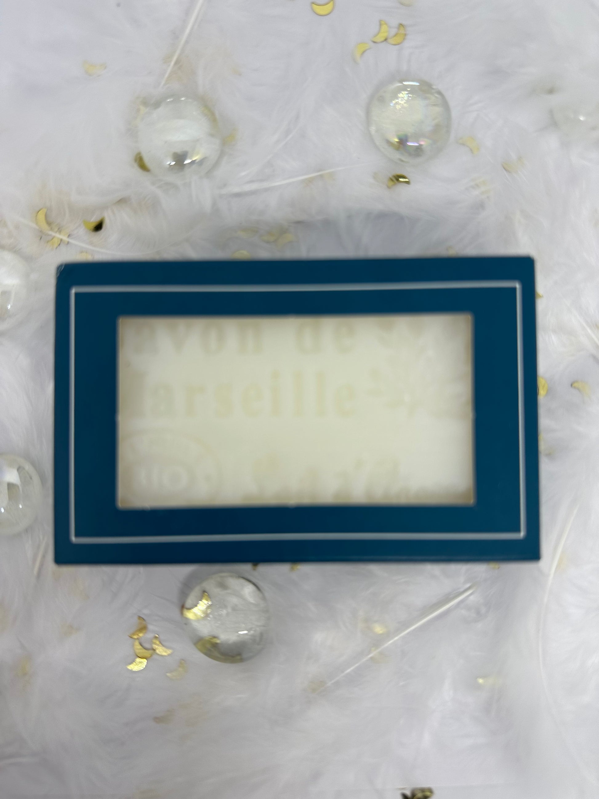 Savon de marseille blanc dans son emballage carton bleu sur fond blanc avec plumes blanches, paillettes dorée et billes transparentes